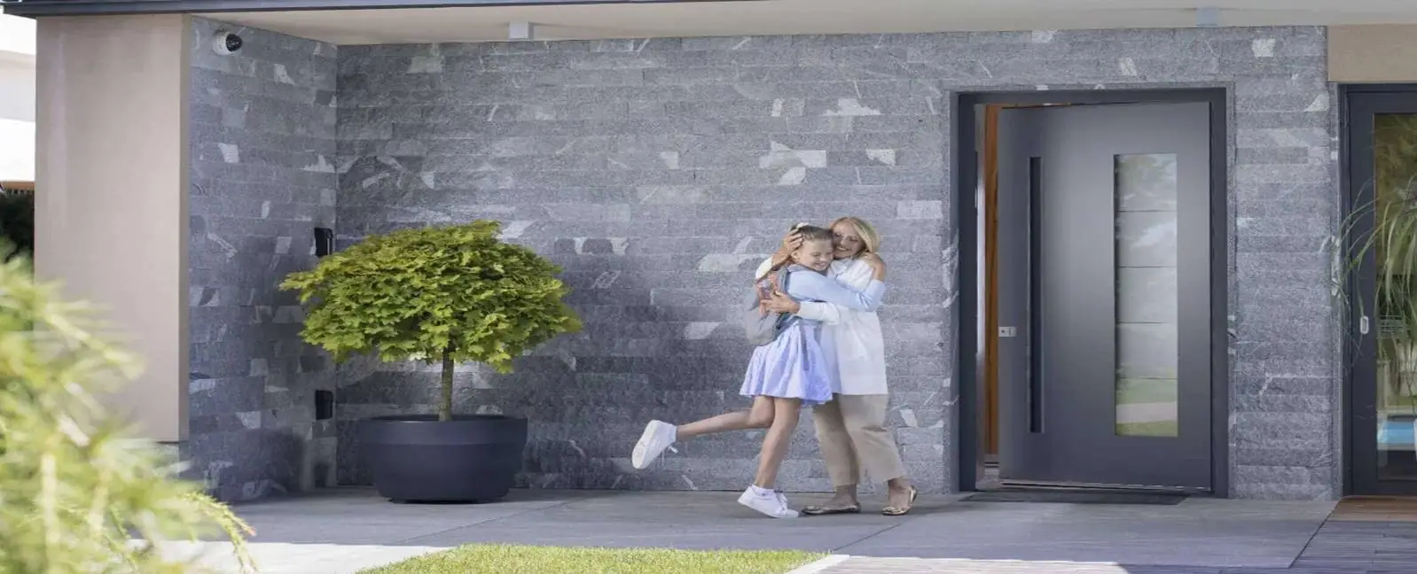 Une mère et sa fille se tenant joyeusement devant une élégante et sécurisée porte d'entrée en aluminium d'une maison contemporaine.